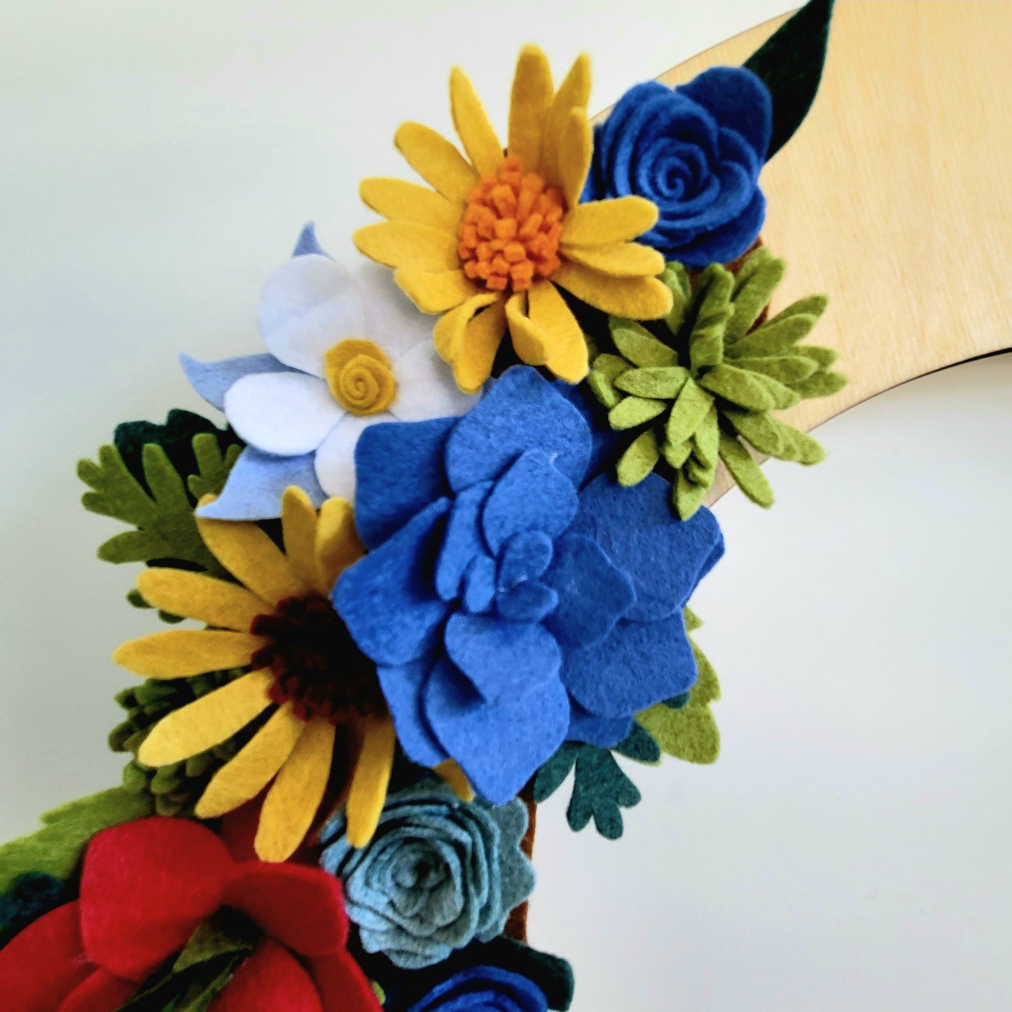A vibrant arrangement of Colorado Alpine Wildflowers is showcased, featuring Eco-Fi felt flowers in various colors. The handmade-in-USA blooms include large blue, yellow, red, and white flowers with green felt leaves. The background is a neutral off-white surface, while a wooden element is partially visible.
