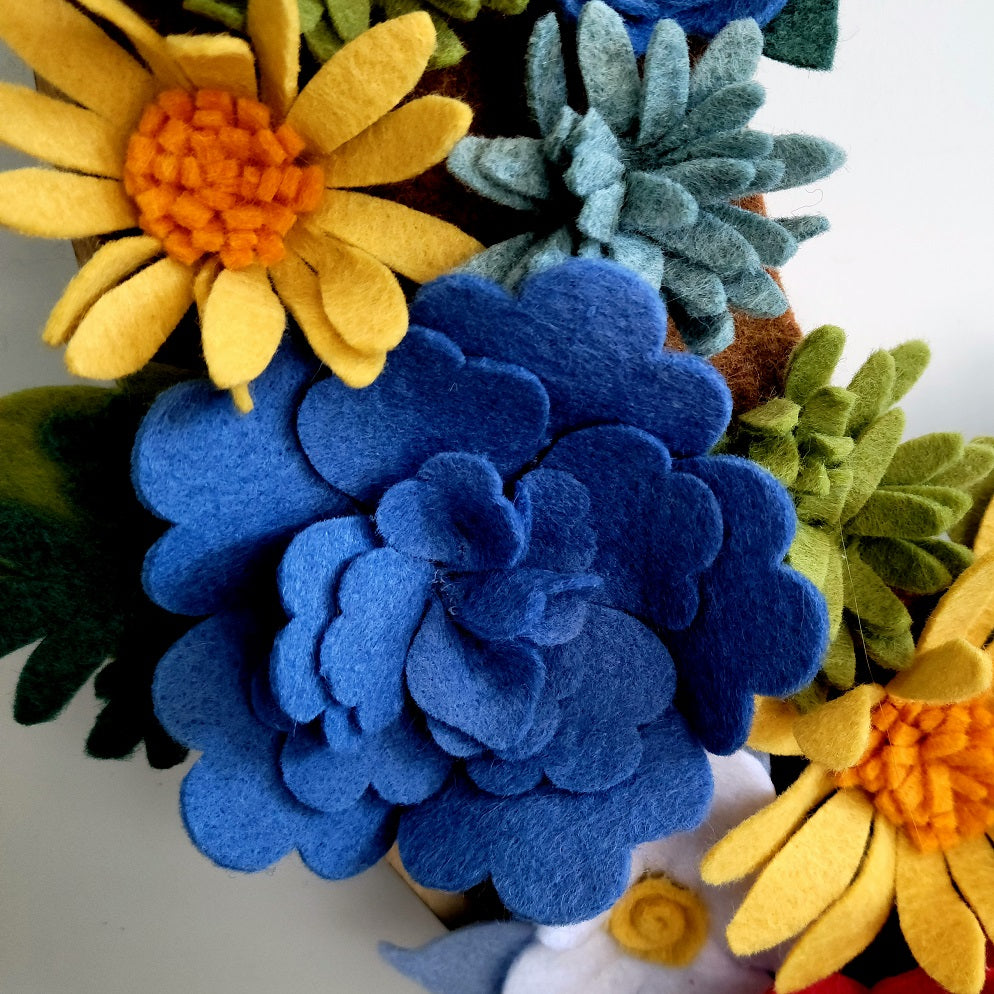 A close-up shot features an assortment of handmade felt flowers from the "Colorado Alpine Wildflowers | Standard Wreath Attachment," crafted in the USA. The focal point is a large blue flower with layered petals, surrounded by yellow and white flowers with orange centers, green foliage, and smaller blue and white blooms made from Eco-Fi felt. The neutral background enhances the vibrant colors.