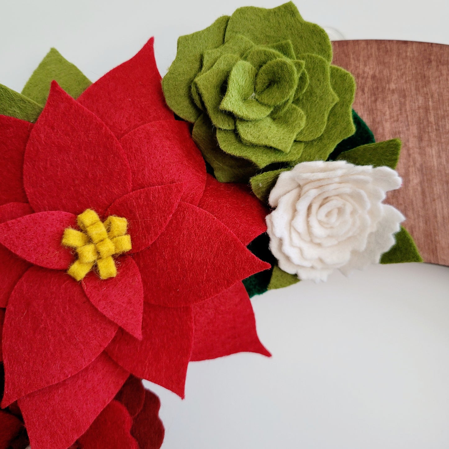 Close-up of felt flowers crafted from variously colored pieces. A large red poinsettia with yellow stamens is accompanied by a green rose and a white rose, each intricately detailed. These flowers, perfect as the Christmas | Standard Wreath Attachment, rest on a wooden surface, highlighting the vibrant colors against the wood grain.
