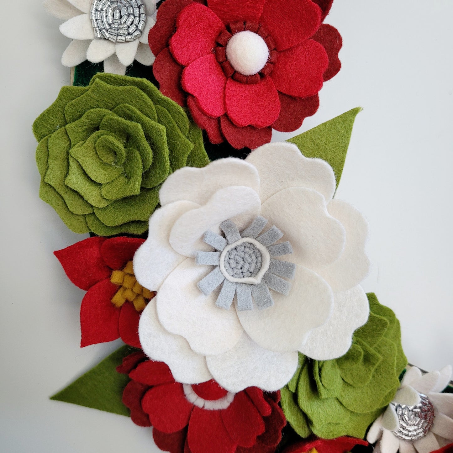 A close-up of handcrafted felt flowers arranged in a cluster. The Christmas | Standard Wreath Attachment, perfect for handmade wreaths, includes white and red poinsettias along with green petals shaped like succulents in various designs. Some flowers are adorned with decorative fabric buttons in the center, showcasing detailed layers and intricate stitching.