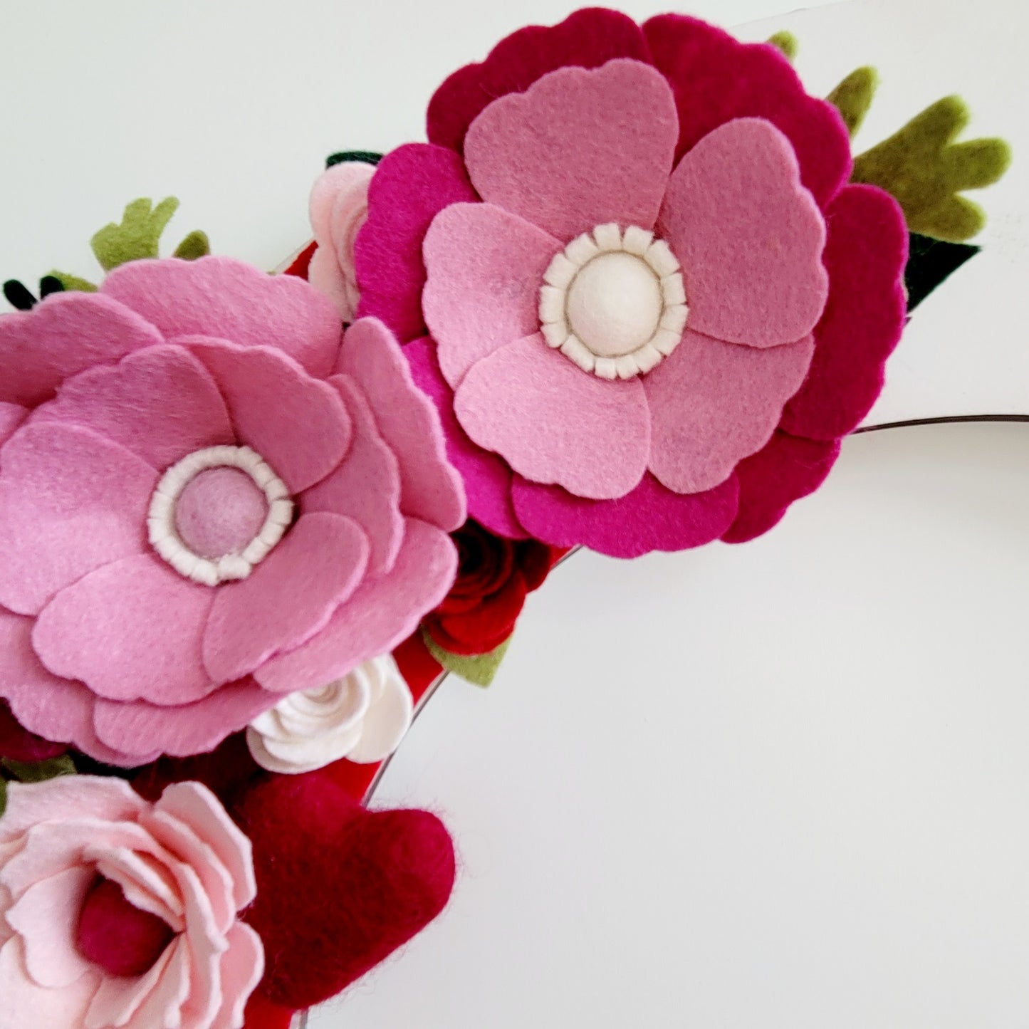 A handmade Valentine’s headband adorned with pink and dark pink Eco-Fi felt flowers of varying sizes, each featuring a white or light pink center, is shown against a plain white background. Small green felt leaves are interspersed among the flowers for a touch of contrast.