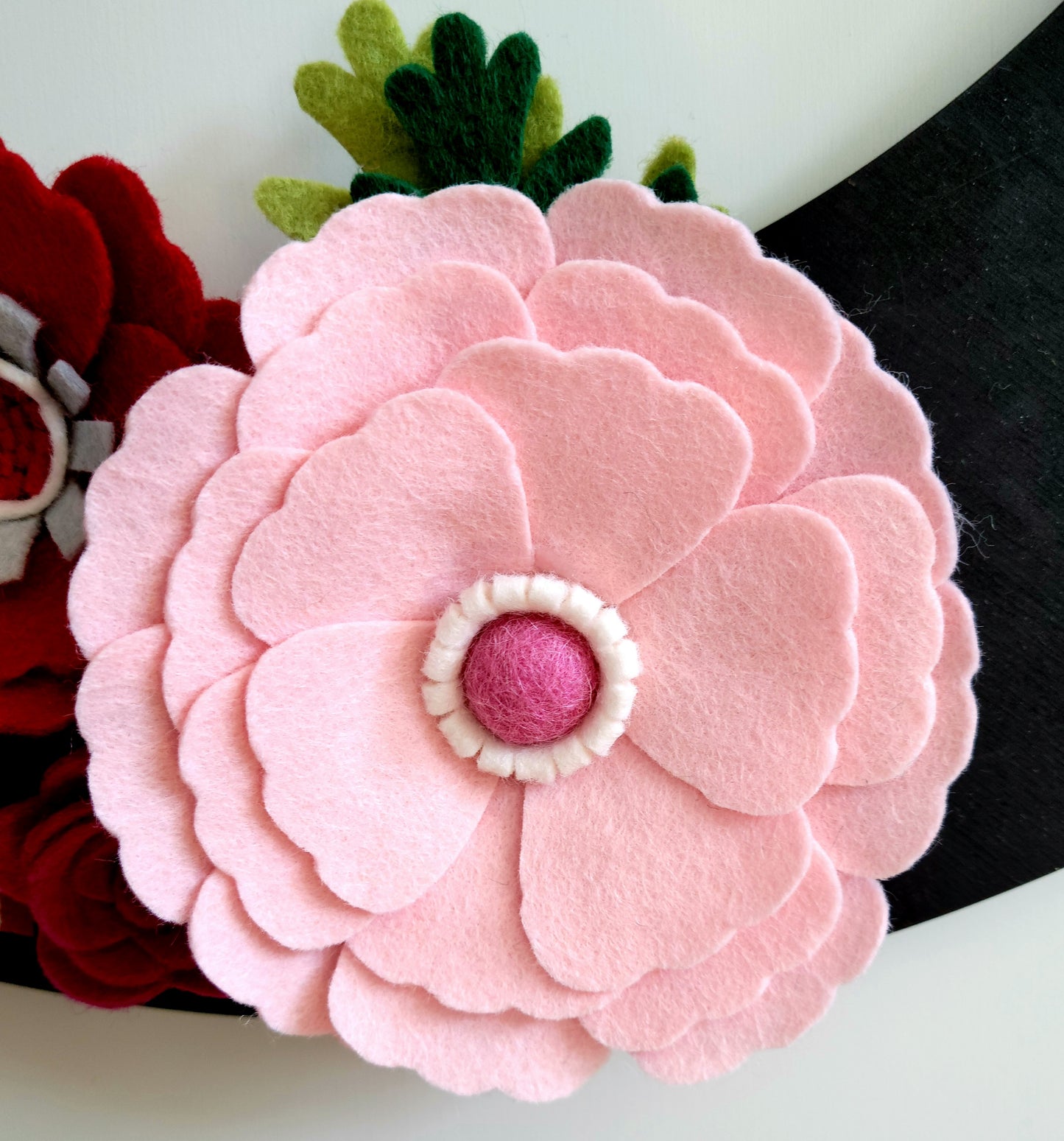 A close-up of a handmade felt flower features soft pink petals, a red center, and white detailing around the center. Green Eco-Fi felt leaves are visible in the background. The flower and leaves are attached to the edge of a black object, indicating it could be part of the "Valentines | Standard Wreath Attachment." The craftsmanship highlights the texture and layers of the felt beautifully.
