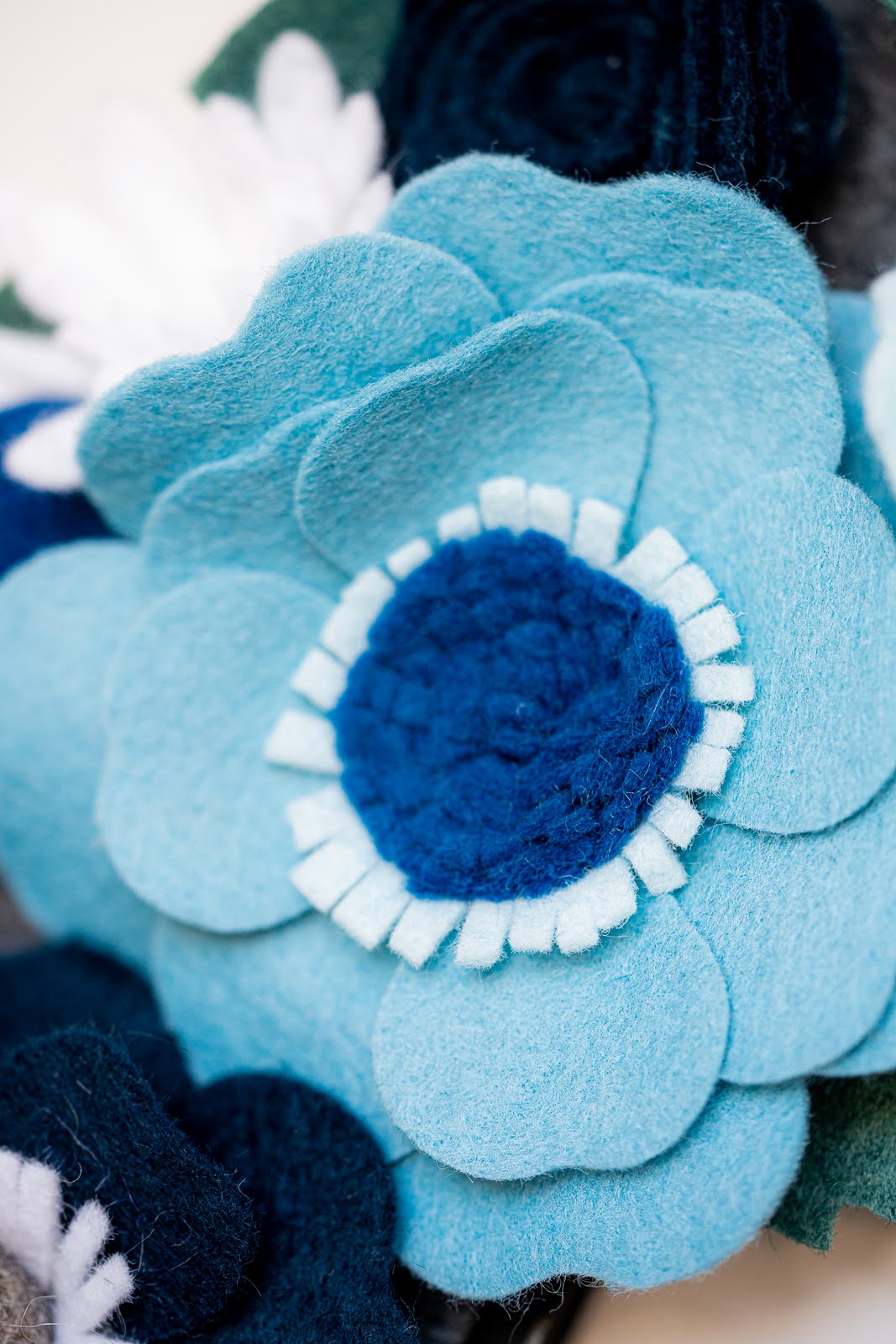 Close-up of the Winter Standard Wreath Attachment, a decorative flower crafted from various shades of blue Eco-Fi felt fabric. This handmade flower features layered petals with a light blue outer layer and a darker blue center, surrounded by fringed light blue felt. The background includes hints of dark blue and white felt details, making it perfect for enhancing any wreath.