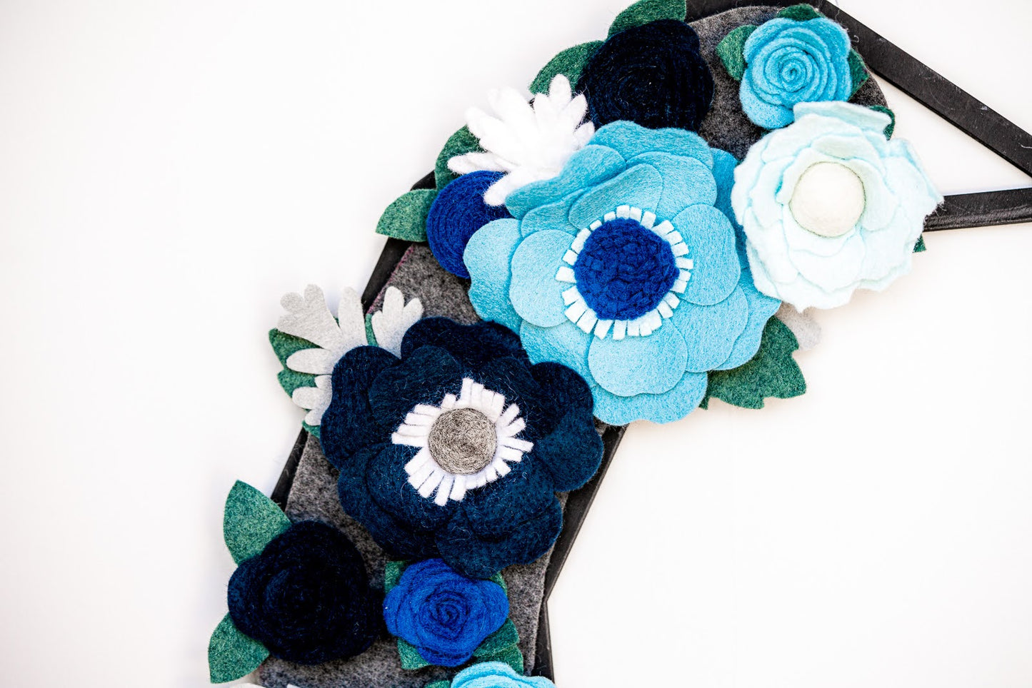 A close-up of the Winter | Standard Wreath Attachment on a gray background. The handmade arrangement showcases large, intricately crafted flowers in shades of blue, white, and gray. Green Eco-Fi felt leaves are interspersed among the flowers, enhancing the visual appeal of the design.