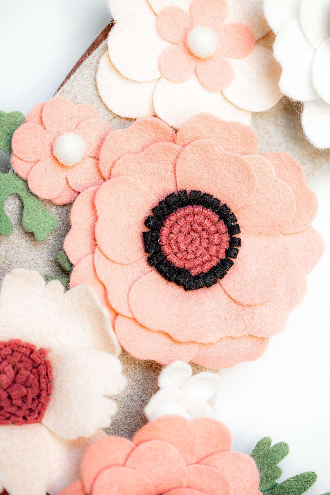 Close-up image of the Spring | Standard Wreath Attachment showcasing handmade felt flowers in various colors. Crafted from Eco-Fi felt, the flowers feature petals in soft pinks, peaches, and whites with detailed centers in shades of red and black. Green felt leaves offer a complementary background, while the texture and layering add depth and dimension.