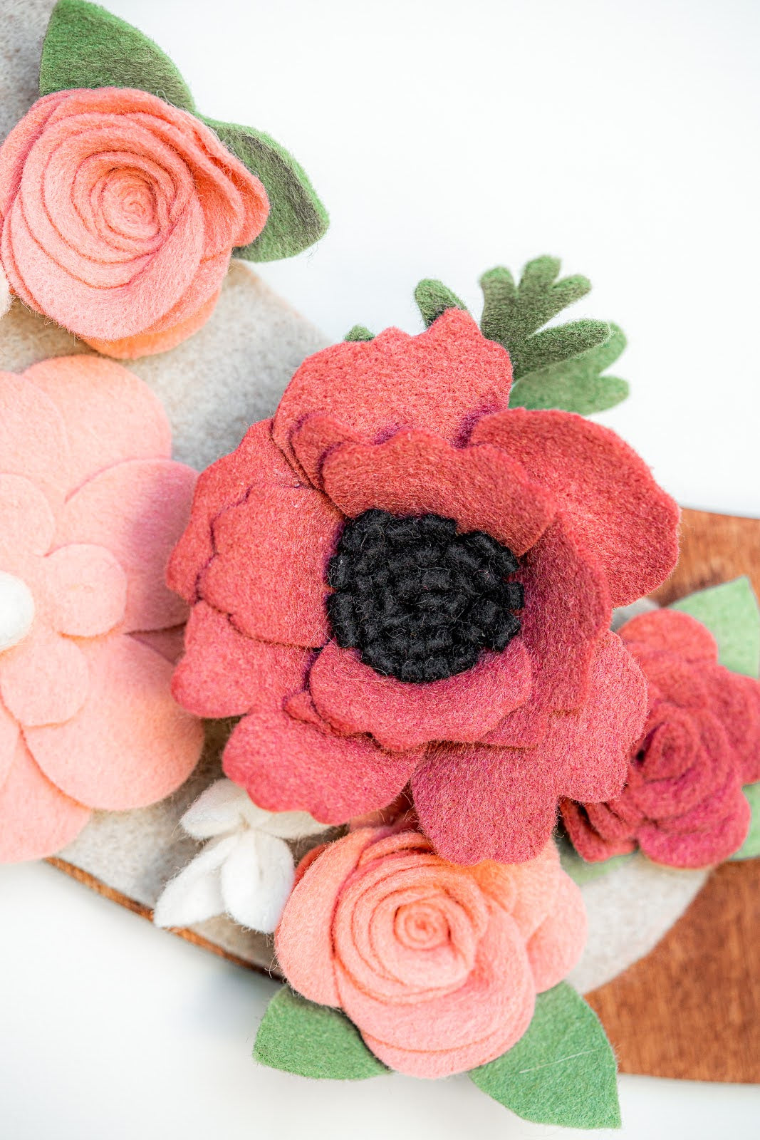 A detailed close-up of the "Spring | Standard Wreath Attachment" featuring a flower arrangement made from recycled materials like Eco-Fi felt. The centerpiece is a large red poppy with a dark center, surrounded by smaller pink and peach roses. Green felt leaves and buds are interspersed among the flowers, all set against a neutral background.