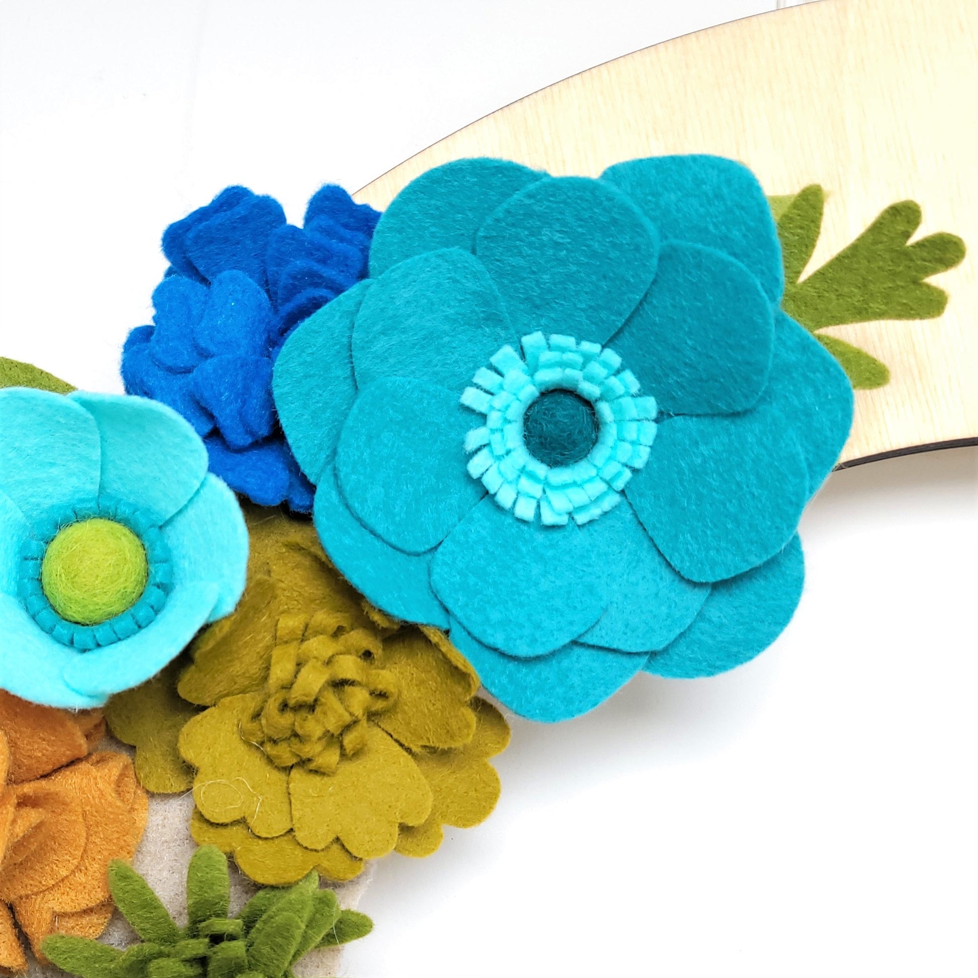 A close-up image features a 'Save the Sea Turtles | Standard Wreath Attachment,' a wooden wreath adorned with vibrant felt flowers. Handmade in the USA, these flowers display shades of turquoise, blue, light blue, olive green, and tan, highlighting detailed petals and centers. The minimal background emphasizes the colorful and intricate craftsmanship of the felt flowers.