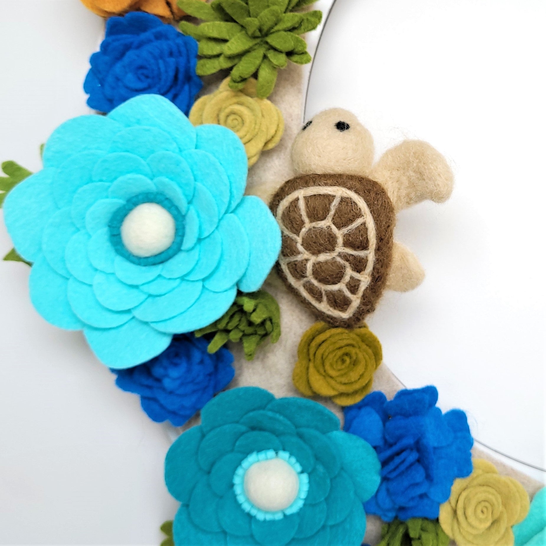 A close-up of the "Save the Sea Turtles" Standard Wreath Attachment showcases vibrant felt flowers in predominantly blue and teal hues, accented with yellow and green. A charming brown felt sea turtle with a beige shell pattern nestles among the flowers, adding a cute touch to this handmade-in-USA decorative piece.